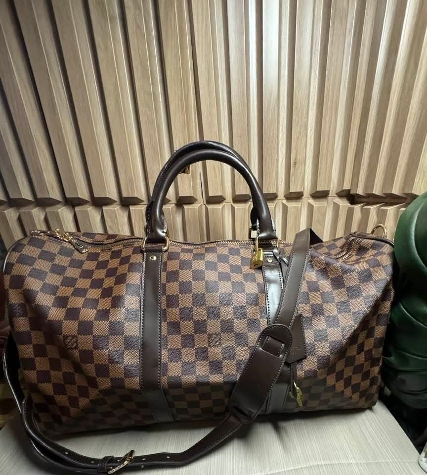 Louis Vuitton Damier Ebene Keepall Bandouliere (Category: Luxury Bags & Wallets) located in Manila. Submitted by goodbuying_items0211 (Listing ID 1467969207, Image 1). Description: - Louis Vuitton travel duffel in classic Damier Ebene canvas - Dark brown leather trim and polished gold-tone hardware - Spacious red fabric interior featuring a convenient zip pocket - Includes matching shoulder strap, padlock, and keys - Clean condition inside and ou