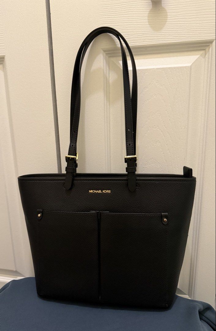 Michael Kors Black Jet Set Medium Pocket Tote Bag (Category: Tote Bags) located in Tabuk. Submitted by farawayluxe (Listing ID 1468005149, Image 1). Description: Black pebble leather tote bag from Michael Kors with gold-tone hardware. Double shoulder straps with adjustable buckle details. Front features two open slip pockets and gold metal lettering. Main compartment zips closed. Interior lined with dark patterned fabric and branding throughout. 