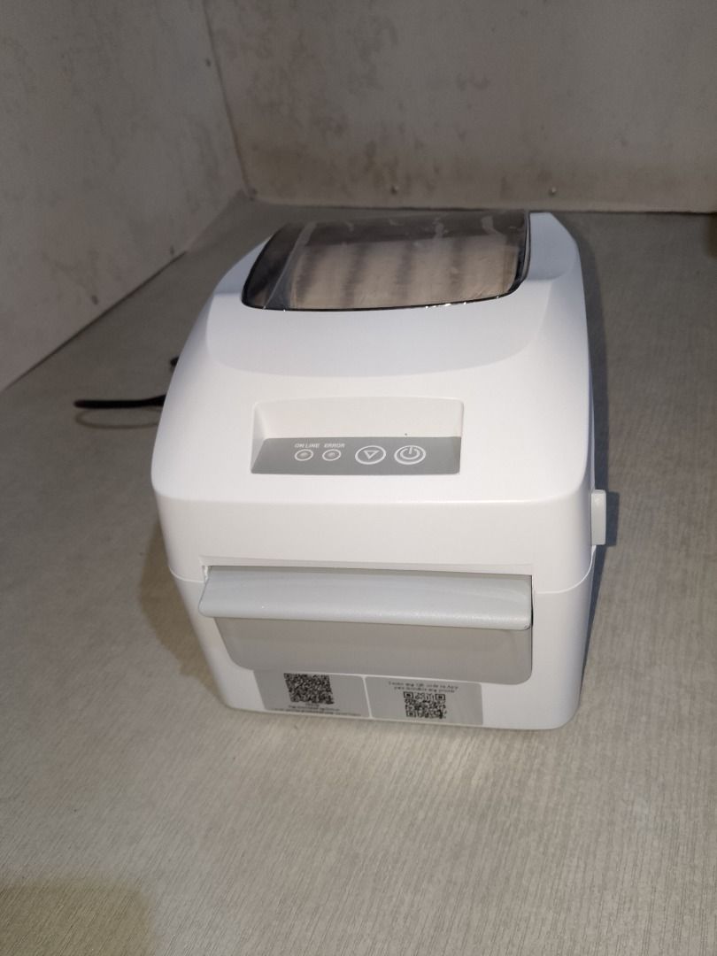 Thermal Label Printer (Category: Printers, Scanners & Copiers) located in Baliuag. Submitted by daisy0215 (Listing ID 1467967656, Image 2). Description: - Sleek white desktop label printer with a space-saving compact footprint - Clear top window lets you easily monitor remaining paper roll - Convenient front control buttons for power and feed - Comes complete with power cable and handy QR code stickers near the bottom edge - Ideal for home office