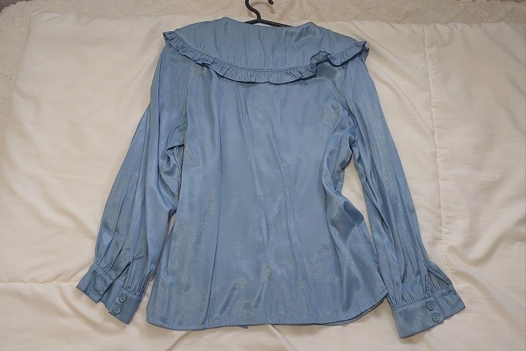 Bench Blue Long Sleeve With Ruffle Collar (Category: Longsleeves) located in Pasig City. Submitted by salazaralona (Listing ID 1468064413, Image 2). Description: - Color: Light blue - Design: Large ruffled collar with front button closure - Sleeves: Long sleeves with buttoned cuffs - Fabric: Soft material with a subtle sheen - Style: Versatile piece, great for casual wear or dressing up.