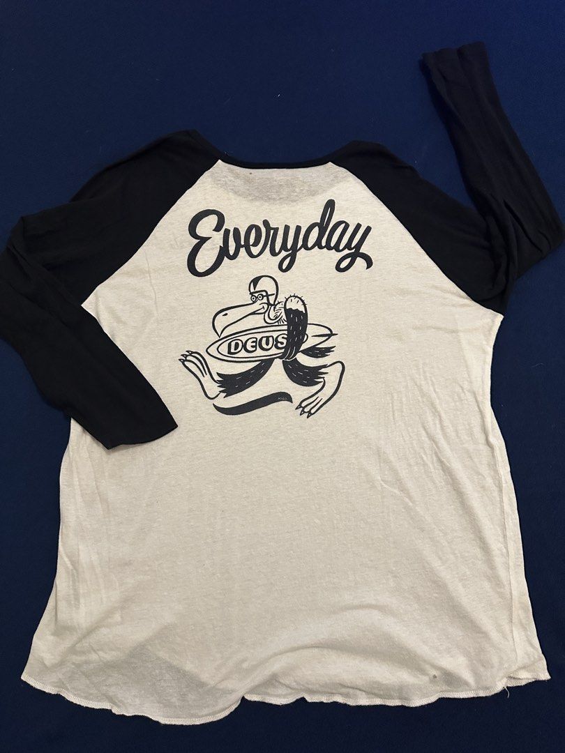 Deus ex Machina (Category: Activewear) located in Quezon City. Submitted by mancave_stuff (Listing ID 1468155154, Image 2). Description: Deus ex Machina 3/4 long sleeves Size XL (W22” x L25”) • Style: Raglan-sleeve baseball shirt with contrasting dark sleeves and a light body. • Graphic: A custom illustration with script text reading “Everyday” and a surfboard graphic displaying the DEUS name, associated with the lifestyle and moto-surf brand Deu