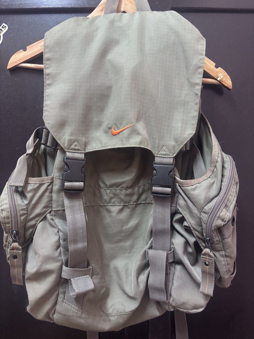 Vintage Nike Olive Green Rucksack Backpack (Category: Backpacks) located in Quezon. Submitted by vintageshorks (Listing ID 1468219120, Image 1). Description: - Color: Olive green with orange embroidered Nike swoosh logo - Material: Durable ripstop fabric - Closure: Flap top with dual quick-release plastic buckles - Storage: Multiple exterior side zip pockets and interior mesh pocket - Comfort: Padded mesh back panel and adjustable shoulder straps