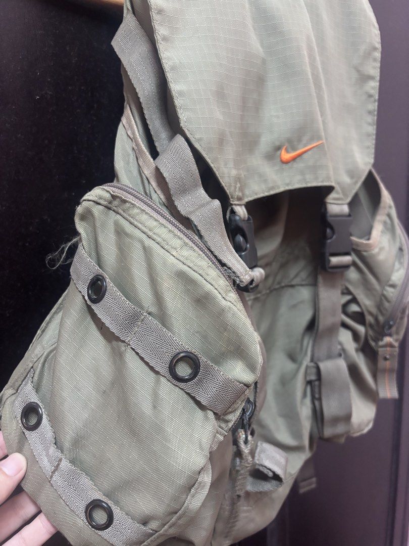 Vintage Nike Olive Green Rucksack Backpack (Category: Backpacks) located in Quezon. Submitted by vintageshorks (Listing ID 1468219120, Image 2). Description: - Color: Olive green with orange embroidered Nike swoosh logo - Material: Durable ripstop fabric - Closure: Flap top with dual quick-release plastic buckles - Storage: Multiple exterior side zip pockets and interior mesh pocket - Comfort: Padded mesh back panel and adjustable shoulder straps