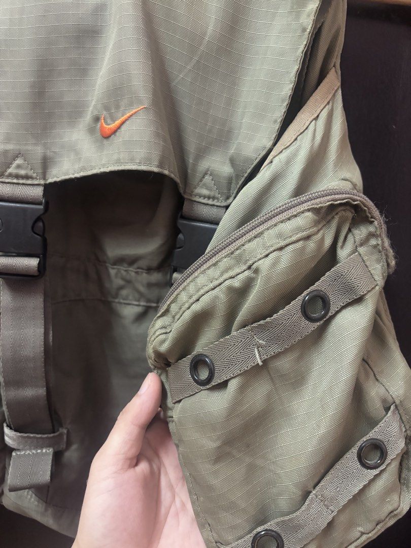 Vintage Nike Olive Green Rucksack Backpack (Category: Backpacks) located in Quezon. Submitted by vintageshorks (Listing ID 1468219120, Image 3). Description: - Color: Olive green with orange embroidered Nike swoosh logo - Material: Durable ripstop fabric - Closure: Flap top with dual quick-release plastic buckles - Storage: Multiple exterior side zip pockets and interior mesh pocket - Comfort: Padded mesh back panel and adjustable shoulder straps