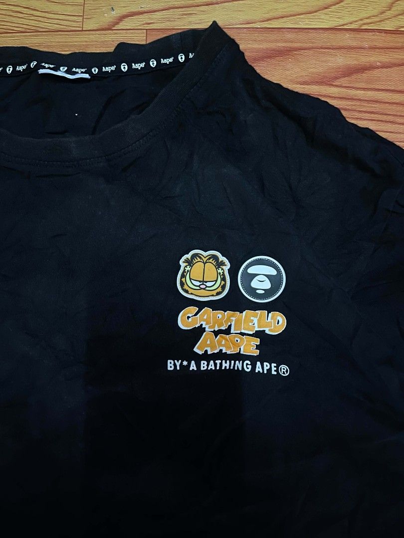 Aape x Garfield Black T-Shirt (Category: Tshirts & Polo Shirts ) located in Ozamiz City. Submitted by tripft.vonde (Listing ID 1468312263, Image 2). Description: Black short sleeve t-shirt from the Aape by A Bathing Ape and Garfield collaboration. Small Garfield and Aape branding on the left chest. Larger graffiti-style graphic on the back with camouflage fill. Small woven logo tags on the left sleeve hem. Collar tape features repeated Aape text.