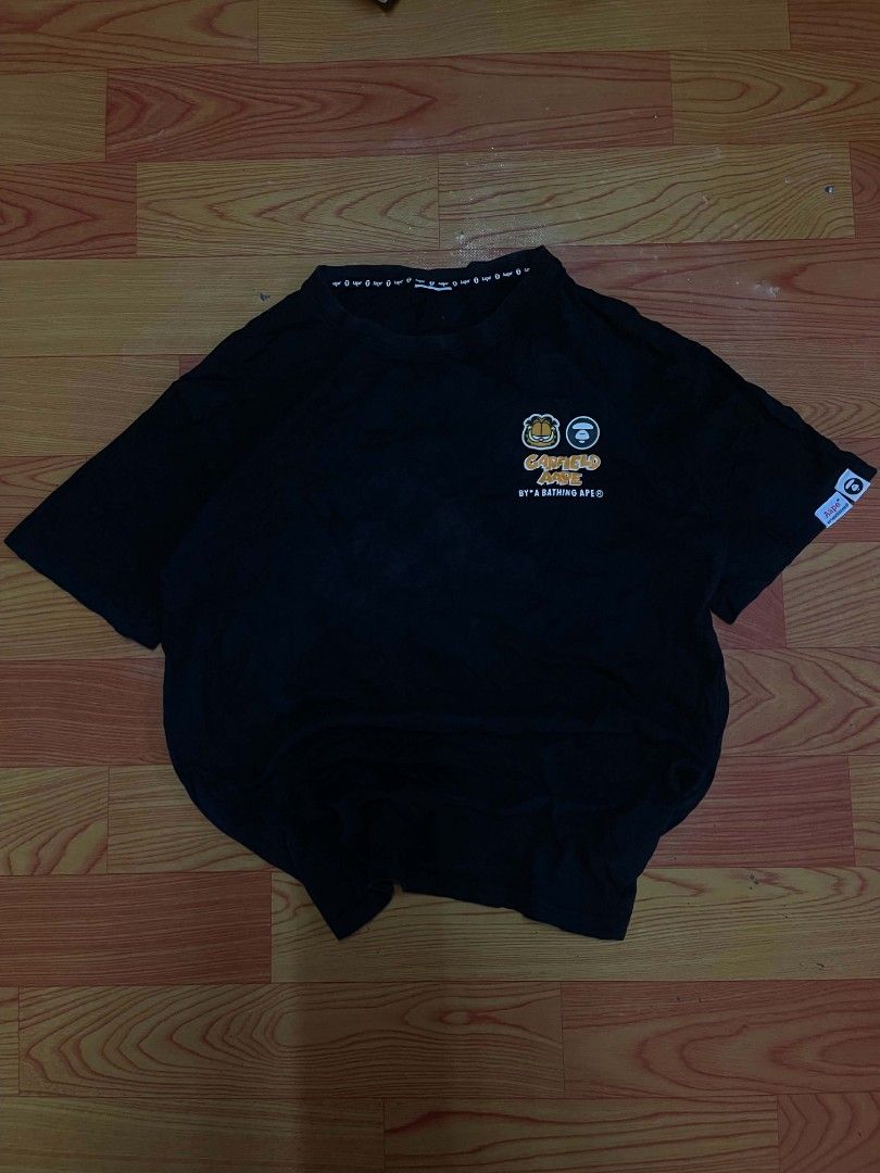 Aape x Garfield Black T-Shirt (Category: Tshirts & Polo Shirts ) located in Ozamiz City. Submitted by tripft.vonde (Listing ID 1468312263, Image 1). Description: Black short sleeve t-shirt from the Aape by A Bathing Ape and Garfield collaboration. Small Garfield and Aape branding on the left chest. Larger graffiti-style graphic on the back with camouflage fill. Small woven logo tags on the left sleeve hem. Collar tape features repeated Aape text.