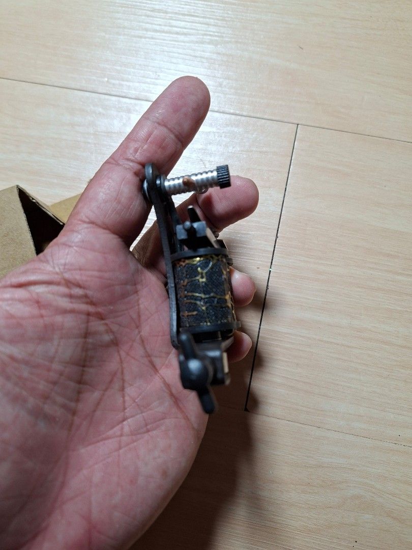 Coil Tattoo Machine Black and Gold (Category: Craft Supplies & Tools) located in Baguio. Submitted by restyrille77846 (Listing ID 1468313817, Image 3). Description: - Color: Black and gold with patterned coil wraps - Setup: Standard setup with binding posts and contact screw - Condition: Brand new, never used, kept in storage - Inclusions: Comes with original cardboard box.