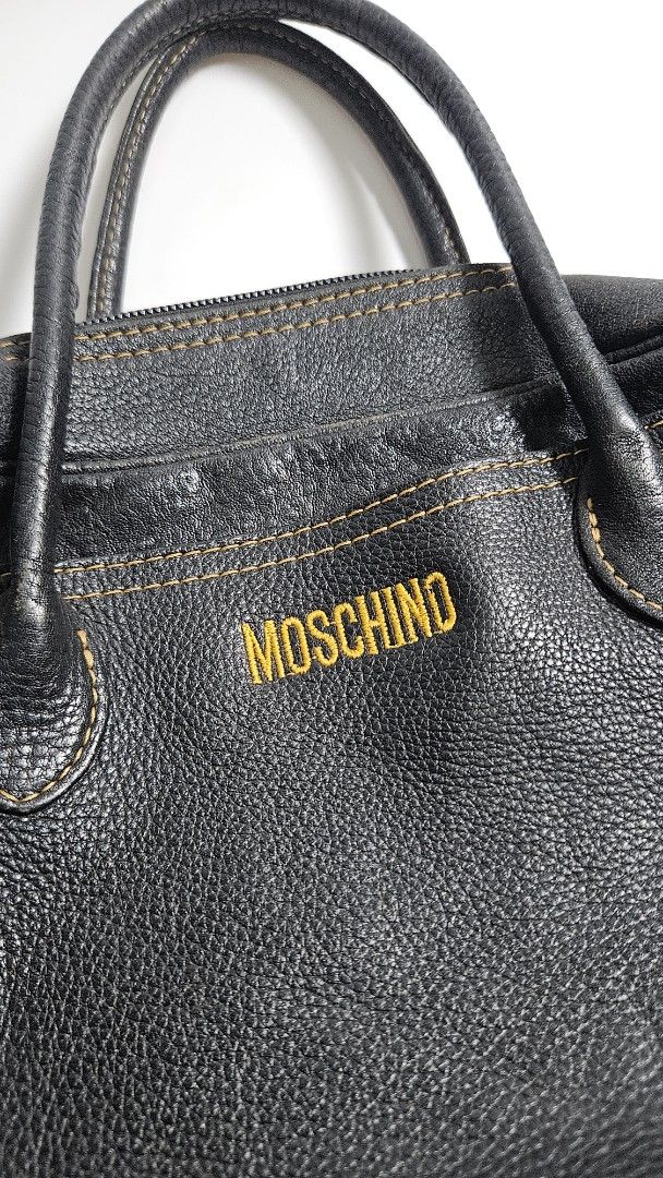 Moschino Black Leather Handbag (Category: Luxury Bags & Wallets) located in Alfonso. Submitted by jbcamet (Listing ID 1468389444, Image 2). Description: - Brand: Moschino - Material: Black pebble leather with fabric lining - Details: Yellow embroidered front logo, contrasting orange stitching - Hardware: Protective metal feet at the base - Closure: Top zip closure with double top handles - Condition: Classic vintage piece with subtle wear - Size: