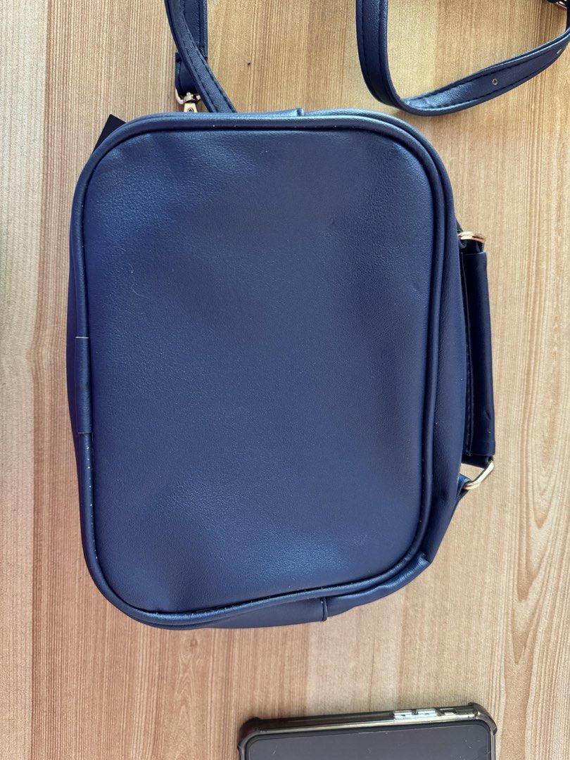 Navy Blue Crossbody Bag with Gold Hardware (Category: Cross-body Bags) located in Valenzuela. Submitted by declutterlangpo (Listing ID 1468295290, Image 2). Description: - Color: Navy Blue - Material: Smooth faux leather with clean stitching - Hardware: Gold-toned with front brand plaque - Condition: Brand new with original tag attached - Strap: Adjustable matching shoulder strap - Design: Rectangular shape with rounded corners.