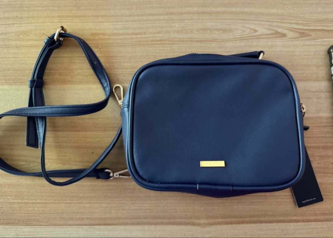 Navy Blue Crossbody Bag with Gold Hardware (Category: Cross-body Bags) located in Valenzuela. Submitted by declutterlangpo (Listing ID 1468295290, Image 1). Description: - Color: Navy Blue - Material: Smooth faux leather with clean stitching - Hardware: Gold-toned with front brand plaque - Condition: Brand new with original tag attached - Strap: Adjustable matching shoulder strap - Design: Rectangular shape with rounded corners.