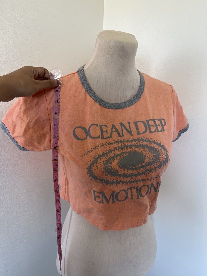 Peach Crop Top (Category: Shirts) located in Mandaluyong City. Submitted by bravedeer.020ad7 (Listing ID 1468366157, Image 2). Description: - Color: Peach with grey ringer neckline and cuffs - Design: 'OCEAN DEEP EMOTIONS' graphic with spiral galaxy center - Fit: Short sleeves, raw hem style, approx. 15 inches from shoulder - Style: Casual crop top, perfect with high-waisted jeans or shorts - Condition: Excellent, well-maintained - Mode of Paymen