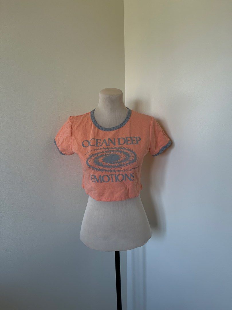 Peach Crop Top (Category: Shirts) located in Mandaluyong City. Submitted by bravedeer.020ad7 (Listing ID 1468366157, Image 1). Description: - Color: Peach with grey ringer neckline and cuffs - Design: 'OCEAN DEEP EMOTIONS' graphic with spiral galaxy center - Fit: Short sleeves, raw hem style, approx. 15 inches from shoulder - Style: Casual crop top, perfect with high-waisted jeans or shorts - Condition: Excellent, well-maintained - Mode of Paymen