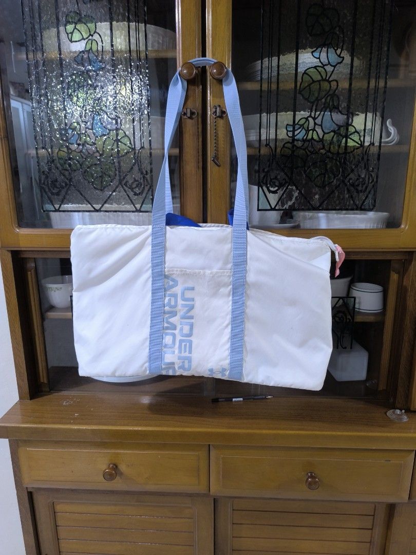Under Armour White Tote Bag (Category: Tote Bags) located in Bay. Submitted by alegnaacir (Listing ID 1468359976, Image 2). Description: - Brand: Under Armour - Color: White with light blue straps and pink zipper pull - Design: Vertical Under Armour branding on the front - Closure: Secure zippered top - Interior: Features UA logo patch and care tags - Material: Durable, lightweight fabric - Condition: Clean overall look - Best for: Gym sessions, 