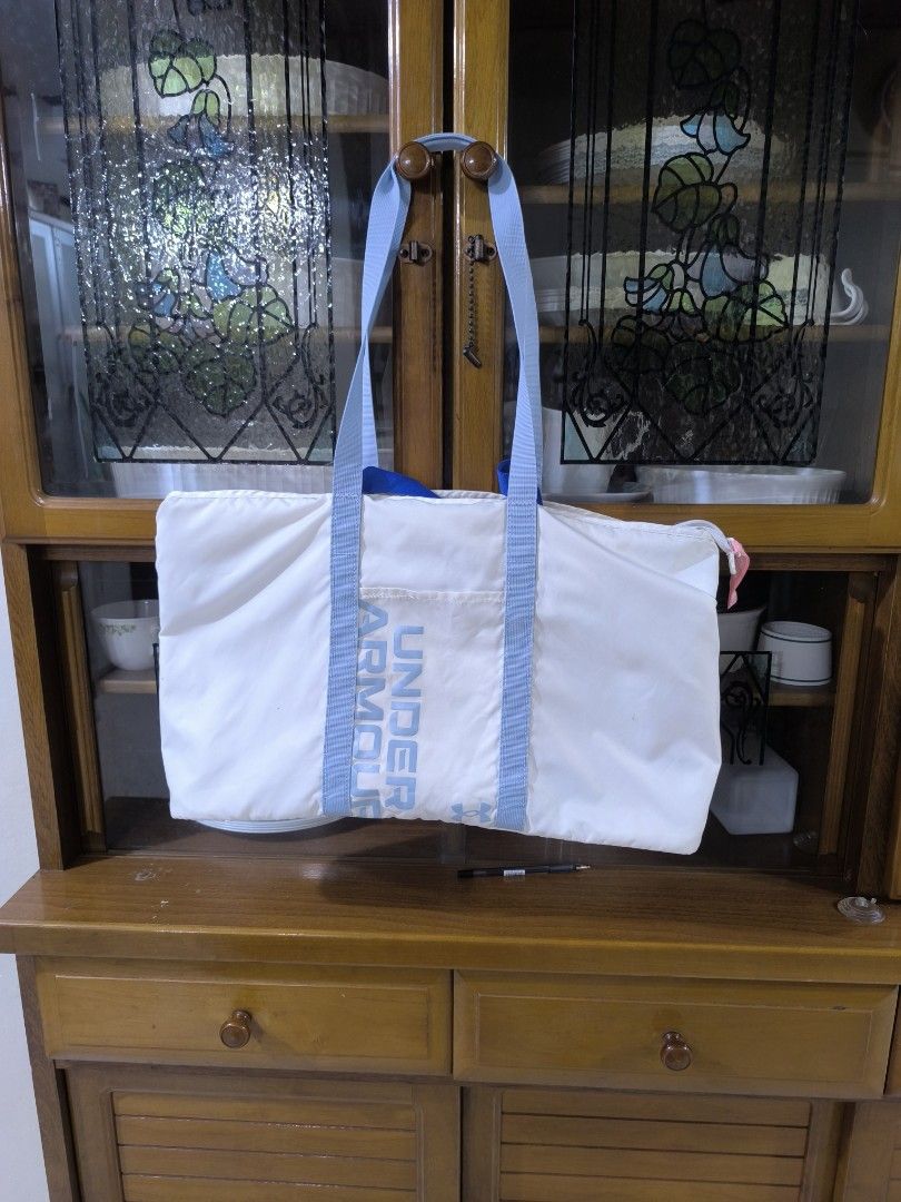 Under Armour White Tote Bag (Category: Tote Bags) located in Bay. Submitted by alegnaacir (Listing ID 1468359976, Image 1). Description: - Brand: Under Armour - Color: White with light blue straps and pink zipper pull - Design: Vertical Under Armour branding on the front - Closure: Secure zippered top - Interior: Features UA logo patch and care tags - Material: Durable, lightweight fabric - Condition: Clean overall look - Best for: Gym sessions, 