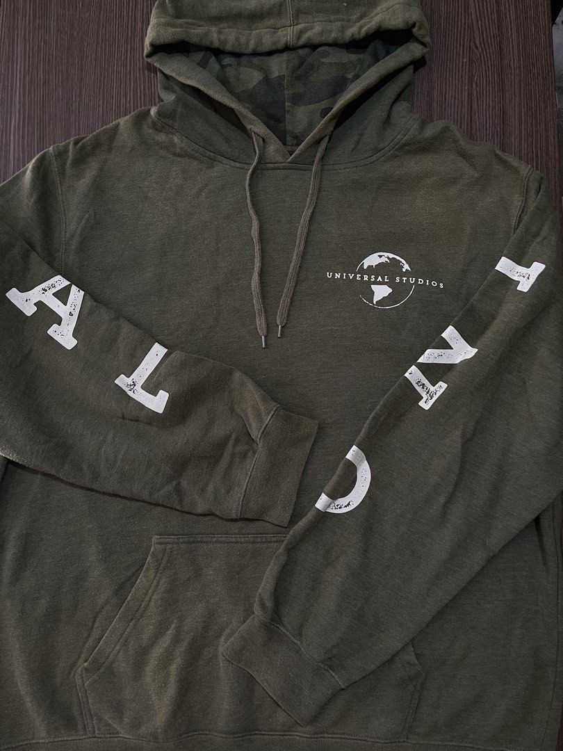 Universal Studios Green Camo Lined Hoodie Size L (Category: Coats, Jackets and Outerwear) located in Manila. Submitted by sidequest2025 (Listing ID 1468396111, Image 2). Description: UNIVERSAL STUDIOS HOODIE (army green) Large on tag, L&W: 22X18 sleeve: 20, shoulder:16 - Brand: Universal Studios - Size: Large (fits true to size) - Color: Olive green with matching camo hood lining - Details: White chest logo, text down sleeves, front kangaroo pock