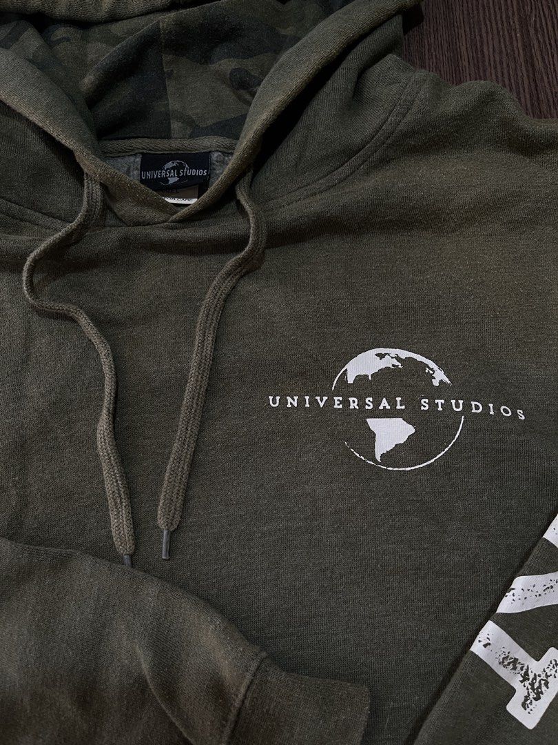 Universal Studios Green Camo Lined Hoodie Size L (Category: Coats, Jackets and Outerwear) located in Manila. Submitted by sidequest2025 (Listing ID 1468396111, Image 3). Description: UNIVERSAL STUDIOS HOODIE (army green) Large on tag, L&W: 22X18 sleeve: 20, shoulder:16 - Brand: Universal Studios - Size: Large (fits true to size) - Color: Olive green with matching camo hood lining - Details: White chest logo, text down sleeves, front kangaroo pock