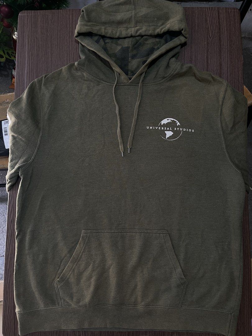 Universal Studios Green Camo Lined Hoodie Size L (Category: Coats, Jackets and Outerwear) located in Manila. Submitted by sidequest2025 (Listing ID 1468396111, Image 1). Description: UNIVERSAL STUDIOS HOODIE (army green) Large on tag, L&W: 22X18 sleeve: 20, shoulder:16 - Brand: Universal Studios - Size: Large (fits true to size) - Color: Olive green with matching camo hood lining - Details: White chest logo, text down sleeves, front kangaroo pock