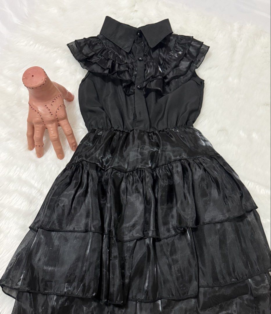 Wednesday Addams Halloween Costume (Category: Babies & Kids Fashion) located in Lemery. Submitted by lovedbyeli (Listing ID 1468376185, Image 2). Description: - Color: Black - Style: Sleeveless mini dress with Wednesday Addams vibe - Details: Layered shoulder ruffles, tiered skirt, collar neckline, and front button closure - Fit: Elasticated waist for easy and comfortable fit - Material: Lightweight, slightly sheer with subtle texture - Inclusion