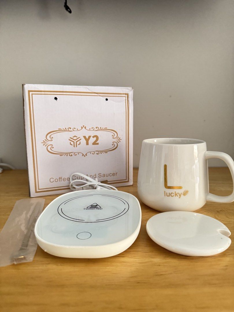Y2 Coffee Cup and Saucer Mug Warmer Set (Category: Coffee & Tea Tableware) located in Mandaluyong City. Submitted by uol77 (Listing ID 1468302136, Image 1). Description: Y2 brand smart coffee cup warmer set. Includes white ceramic mug with letter L and lucky text, matching lid, warming coaster plate, golden spoon, and original box. Keeps drinks warm while working at the desk. Plug-in electric warmer base lights up blue when turned on. Everything 
