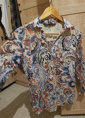 Zara Basic Paisley Print Button-Up Blouse (Category: Blouses) located in Lupao. Submitted by selahthriftedclothing (Listing ID 1468375550, Image 1). Description: - Brand: Zara Basic - Design: White with blue and orange paisley pattern all over - Neckline: V-neck collar - Fabric: Lightweight and comfortable - Versatility: Perfect for casual daily wear or office styling - Pairing: Easy to match with denim or trousers - Siting: Standard sizing.