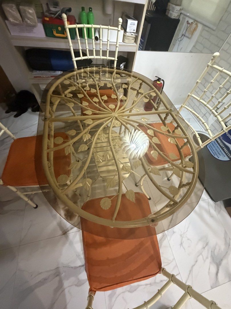4 seater glass top dining set (Category: Tables & Sets) located in Manila. Submitted by noblebird-f4dd (Listing ID 1468579238, Image 3). Description: - Elegant oval dining set with a tinted glass top and decorative leaf/branch metal frame - Includes 4 matching chairs with comfortable orange cushioned seats - Features stylish cream-colored metal legs and classic spindle backrests - Condition: Clean and structurally solid - Perfect for updating you