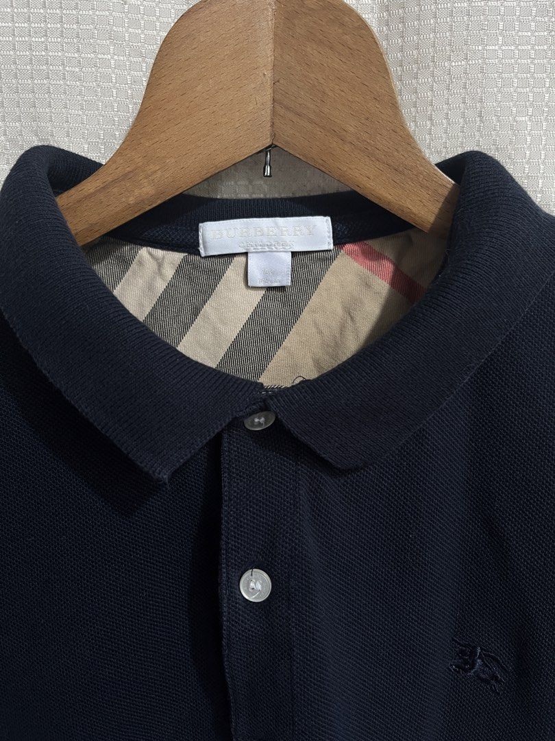 Authentic Burberry Polo Shirt (Category: Shirts) located in Davao. Submitted by aelane.listings (Listing ID 1468499976, Image 3). Description: Color: Black or dark blue Size: 14 Youth on tag, may fit XS-S adult Measurements: 16 1/2in x 25in - Iconic nova check pattern on inside collar and placket - Small embroidered equestrian knight on left chest - Two pearl-tone front buttons Short sleeves, ribbed cuffs and collar Condition: Well-used Clean dar