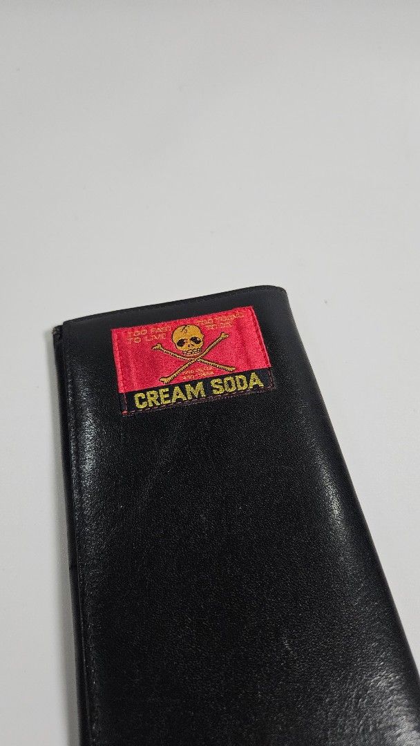 Cream Soda Black Leather Long Wallet (Category: Wallets & Card Holders) located in Alfonso. Submitted by jbcamet (Listing ID 1468652110, Image 2). Description: - Brand: Cream Soda - Material: Smooth black leather - Design: Red exterior label with skull & crossbones and "Too Fast To Live Too Young To Die" text - Interior: Gold skull print with multiple card slots and cash compartments - Condition: Clean stitching and well-maintained - Perfect for 