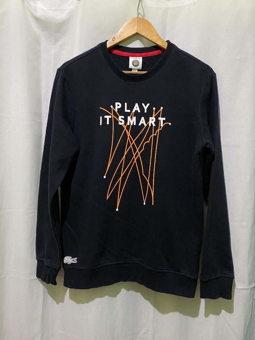Lacoste Play It Smart Black Sweatshirt (Category: Coats, Jackets and Outerwear) located in Quezon City. Submitted by elihur.tuazon (Listing ID 1468593097, Image 2). Description: - Brand: Lacoste - Color: Dark navy blue with red interior collar lining - Size: Large - Design: Large 'Play It Smart' front graphic print with abstract orange lines - Details: Iconic embroidered crocodile logo near the bottom hem - Finish: Ribbed crew neck, cuffs, and he