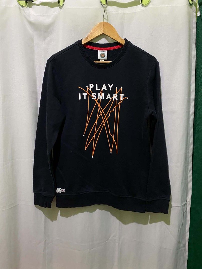 Lacoste Play It Smart Black Sweatshirt (Category: Coats, Jackets and Outerwear) located in Quezon City. Submitted by elihur.tuazon (Listing ID 1468593097, Image 1). Description: - Brand: Lacoste - Color: Dark navy blue with red interior collar lining - Size: Large - Design: Large 'Play It Smart' front graphic print with abstract orange lines - Details: Iconic embroidered crocodile logo near the bottom hem - Finish: Ribbed crew neck, cuffs, and he