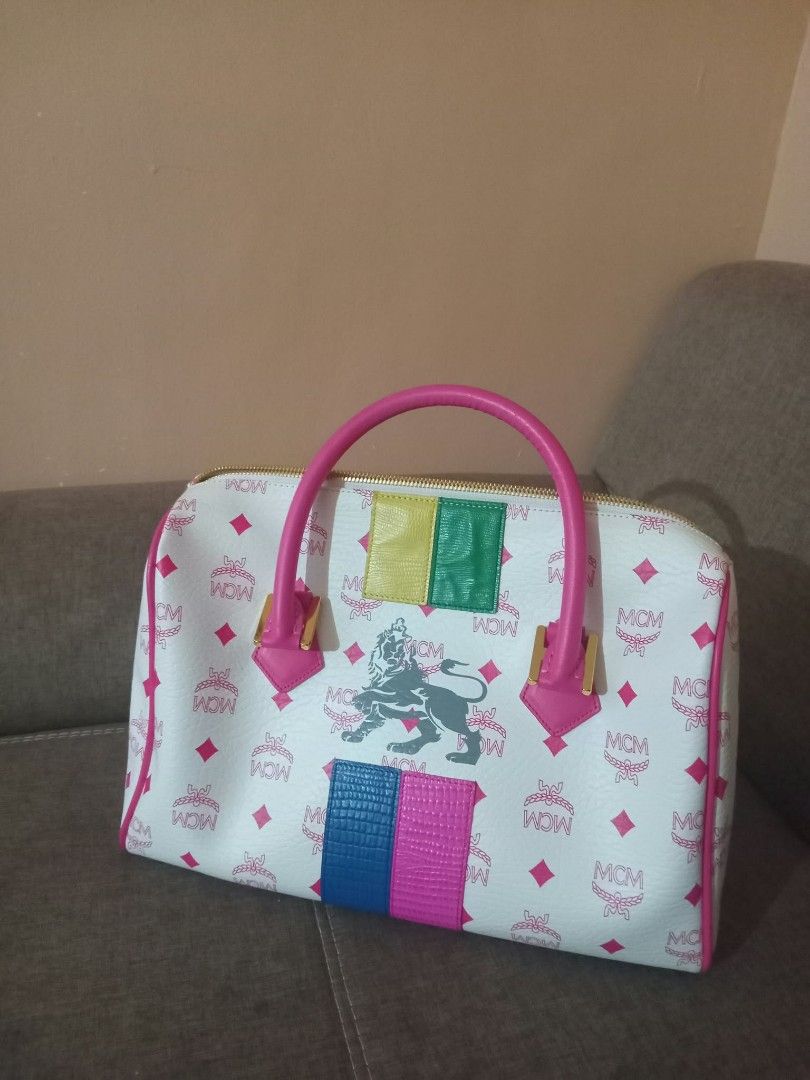 MCM White Pink Monogram Lion Graphic Duffle Handbag (Category: Luxury Bags & Wallets) located in Urdaneta. Submitted by assortedprelovedd (Listing ID 1468585701, Image 1). Description: - Material: White faux leather with pink MCM monogram print - Design: Front lion graphic with vibrant textured leather patches (yellow, green, blue, pink) - Handles & Hardware: Dual pink top handles with gold-tone hardware - Closure: Zip-around with a playful blue 