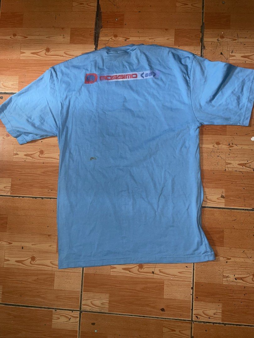 Mossimo 86 Vintage Light Blue T-Shirt (Category: Tshirts & Polo Shirts ) located in Manila. Submitted by joshua_guimeroy. (Listing ID 1468524537, Image 2). Description: - Brand: Mossimo 86 - Color: Light blue - Design: Logo graphic across the chest and matching larger print near the back collar - Details: Short sleeves with a small red brand tag on the left sleeve, standard crew neck fit - Flaw: Small stain visible on the lower back area - Condit