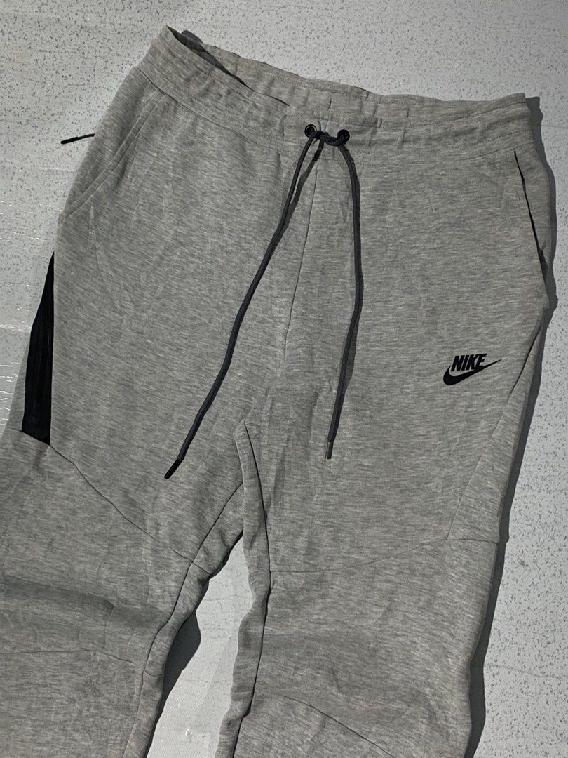 Nike Tech Fleece Joggers Grey (Category: Joggers) located in Manila. Submitted by jeproxthrift (Listing ID 1468626085, Image 2). Description: - Brand: Nike - Color: Grey with black accents - Material: Soft fleece fabric blend - Pockets: Includes a secure zipper pocket on the right leg - Waist: Adjustable drawstring with dark grey cords - Cuffs: Ribbed at the ankles - Branding: Classic embroidered Nike logo on the left thigh.