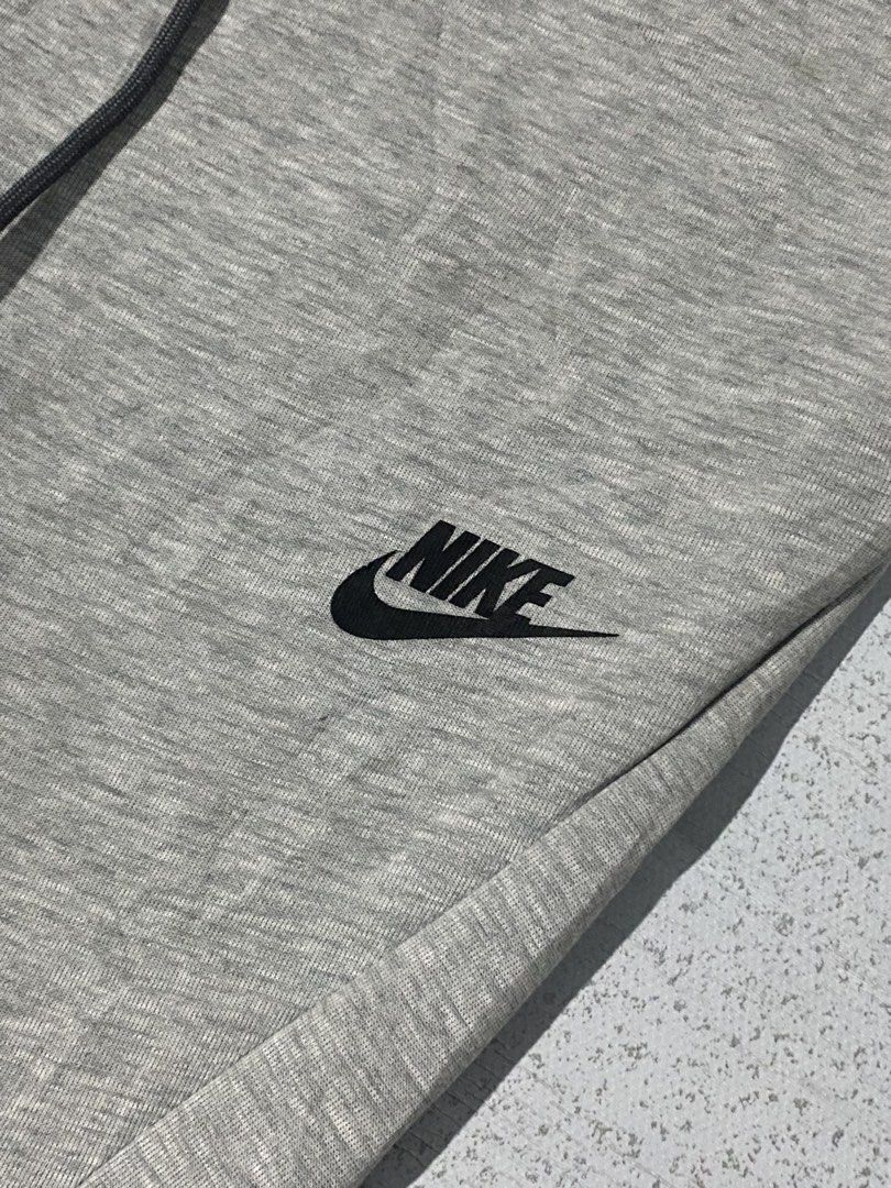 Nike Tech Fleece Joggers Grey (Category: Joggers) located in Manila. Submitted by jeproxthrift (Listing ID 1468626085, Image 3). Description: - Brand: Nike - Color: Grey with black accents - Material: Soft fleece fabric blend - Pockets: Includes a secure zipper pocket on the right leg - Waist: Adjustable drawstring with dark grey cords - Cuffs: Ribbed at the ankles - Branding: Classic embroidered Nike logo on the left thigh.