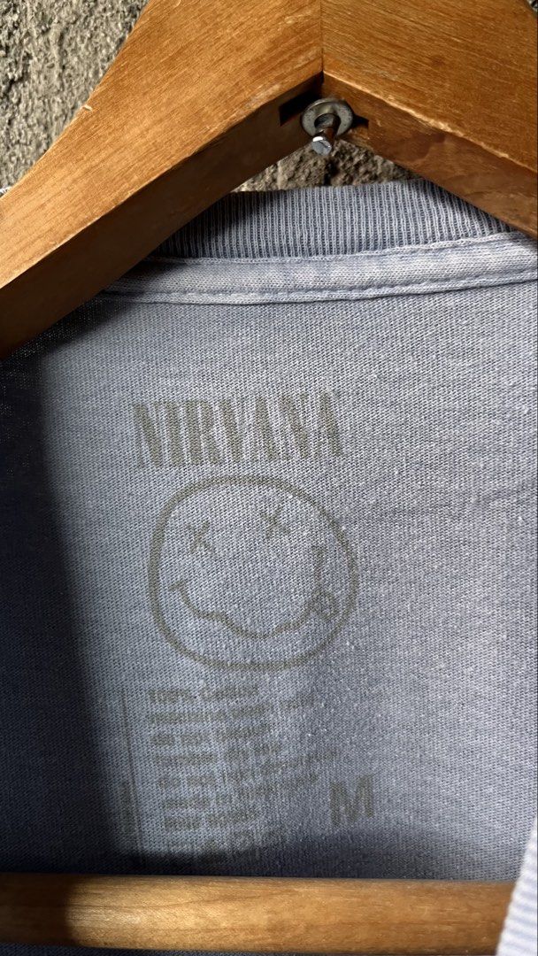 Nirvana Smiley Face Graphic Tee Acid Wash (Category: Tshirts & Polo Shirts ) located in Cabuyao City. Submitted by bulandos (Listing ID 1468636957, Image 2). Description: - Brand: Nirvana - Design: Classic smiley face print - Color/Style: Acid wash faded grey-blue - Size: Medium - Material: 100% cotton, tagless neck label - Condition: Pre-loved with one tiny hole near the bottom hem (shown in close-up photo).