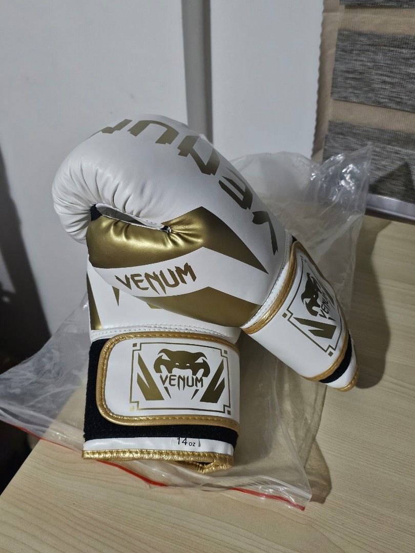 Venum Boxing Gloves 14 oz White Gold (Category: Combat Sports) located in Paranaque City. Submitted by mab.villareal (Listing ID 1468612949, Image 2). Description: Venum boxing gloves in white and gold. Size 14 oz. Velcro strap closure with padded wrist support. Bold Venum logo printed across the front and side. Comes inside clear plastic packaging. Never used, clean condition all around..