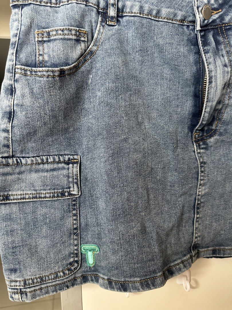 Blue Denim Cargo Skirt with Side Pockets Size M (Category: Skirts) located in Caloocan City. Submitted by be.iiiii (Listing ID 1468828947, Image 2). Description: - Style: Medium wash denim cargo skirt - Details: Front zip and button closure, belt loops, and panel stitching on the back - Pockets: Functional utility side pockets on both hips - Accent: Small turquoise embroidered logo near the bottom left hem - Fit: Classic mid-rise - Pairing: Great