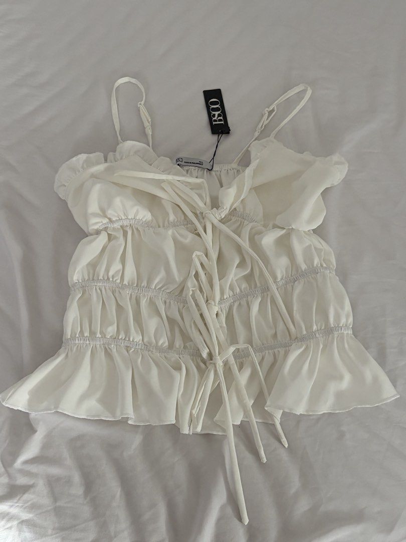 White Spaghetti Strap Top (Category: Sleeveless) located in Las Pinas. Submitted by thriftedfinds_ (Listing ID 1468867882, Image 1). Description: - Brand: BSCO - Condition: Brand new with tags attached - Color: Classic White - Details: Adjustable spaghetti straps, ruffled neckline, smocked tiered body, and front tie strings - Fabric: Lightweight and breathable - Origin: Made in the Philippines - Styling: Perfect to pair with denim shorts or a ski
