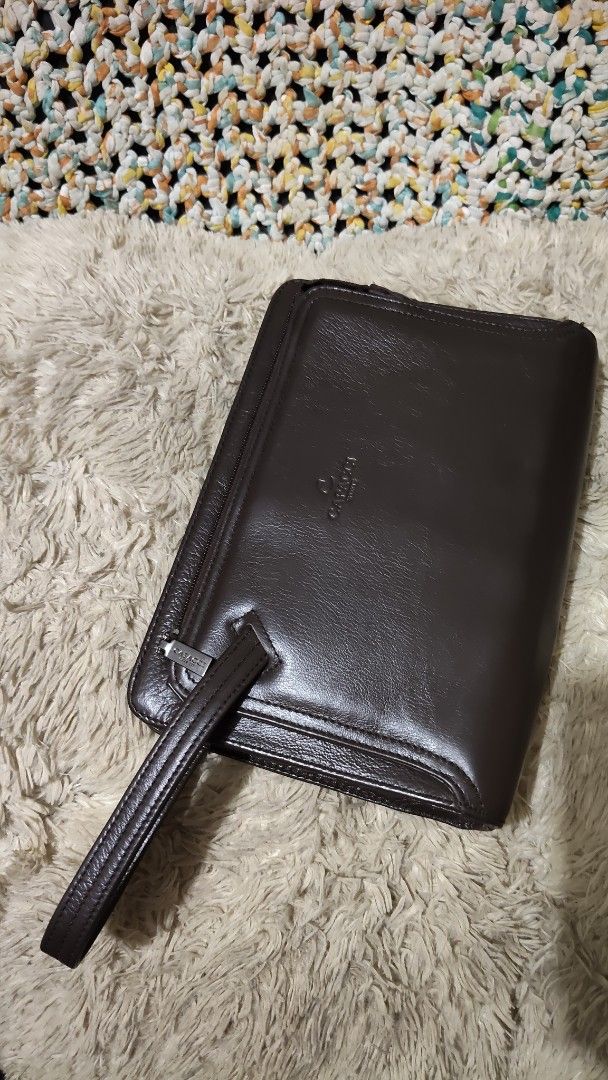Capacci brown leather clutch bag with wrist strap (Category: Clutches) located in Cebu City. Submitted by lumaynojes00490 (Listing ID 1468803188, Image 2). Description: - Brand: Capacci - Color: Deep dark brown with a polished finish - Material: Premium leather with structured rectangular shape - Hardware: Silver-toned with smooth top zipper closure - Features: Attached side wrist strap and embossed front logo.