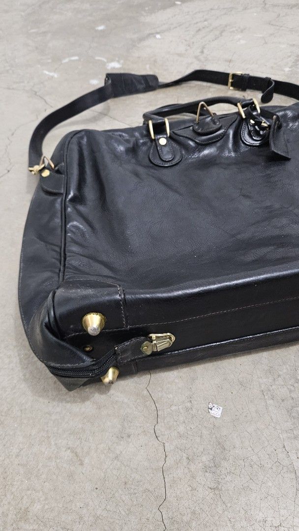 Cole Haan Leather Garment Bag (Category: Briefcases) located in Alfonso. Submitted by jbcamet (Listing ID 1468886069, Image 2). Description: Black leather garment bag from Cole Haan. Made in Italy. Gold tone hardware and protective feet on the bottom. Includes a sturdy shoulder strap with leather pad. Interior lined in beige fabric with multiple zippered pockets, compartments, and a hanger hook. Shows normal signs of wear on the exterior leather 