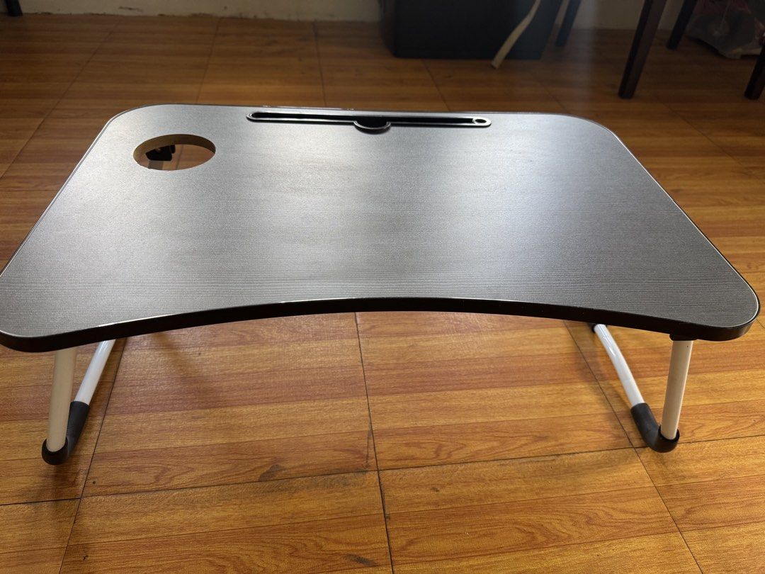 Foldable Laptop Bed Table with Cup Holder (Category: Tables & Sets) located in Mandaluyong City. Submitted by winx3 (Listing ID 1468786814, Image 2). Description: - Type: Portable laptop desk for bed, floor, or couch use - Surface: Dark grey wood-grain with a comfortable curved front edge - Features: Built-in slot for phones/tablets and a round cup holder cutout - Legs: Foldable white metal legs with black anti-slip grips - Condition: Clean surfa
