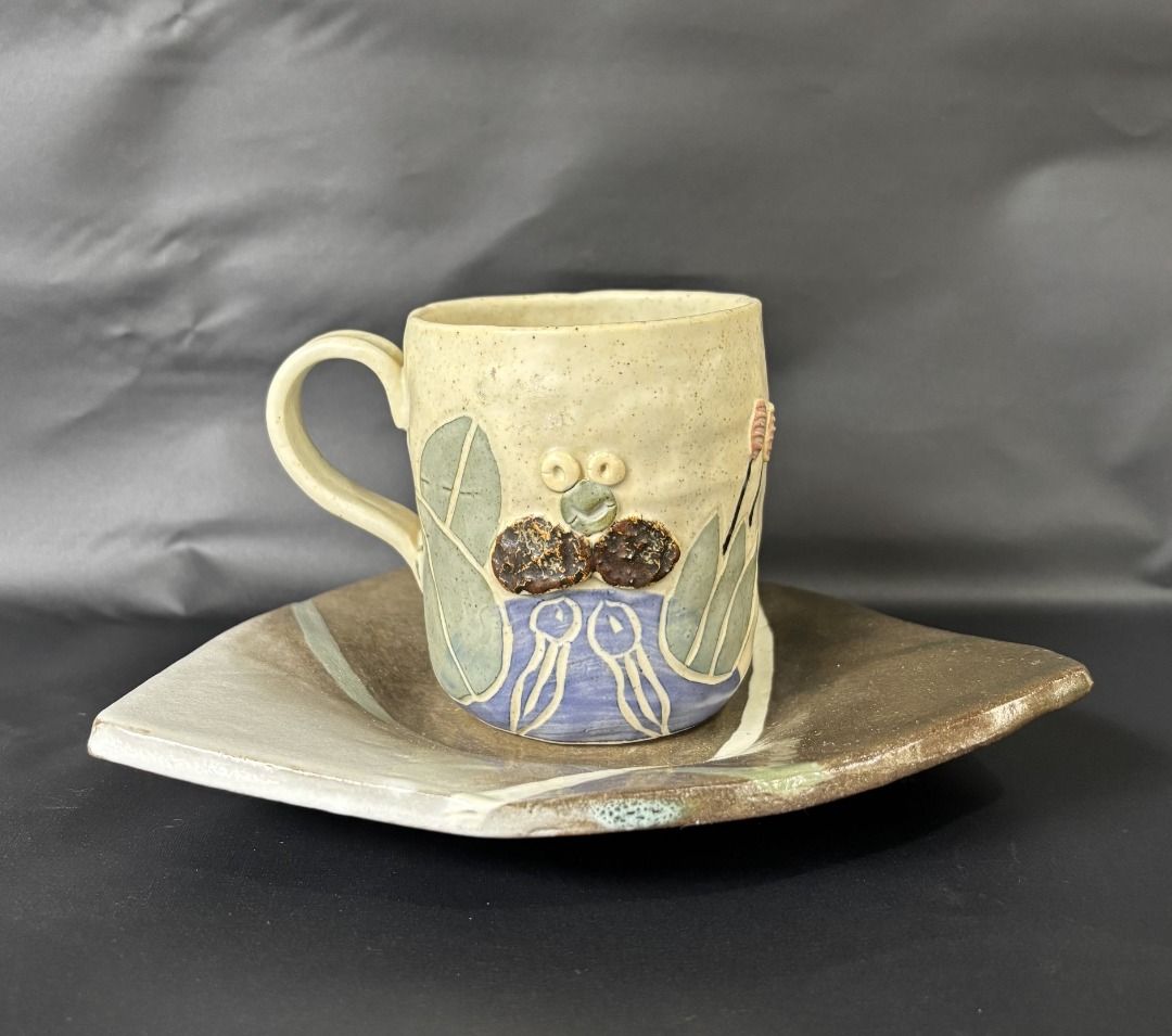 Handmade Ceramic Mug and Triangular Saucer Set (Category: Coffee & Tea Tableware) located in Baao. Submitted by apr1979 (Listing ID 1468785005, Image 1). Description: - Cup: 3.1 x 3.7 inches Triangle Footed Saucer/Trinket: 8 x 1.1 inches - Handmade ceramic mug with a cute raised frog face and plant designs - Glazed in charming beige, blue, and green tones - Comes with a unique triangular saucer featuring earthy brown and grey glazes with painted 