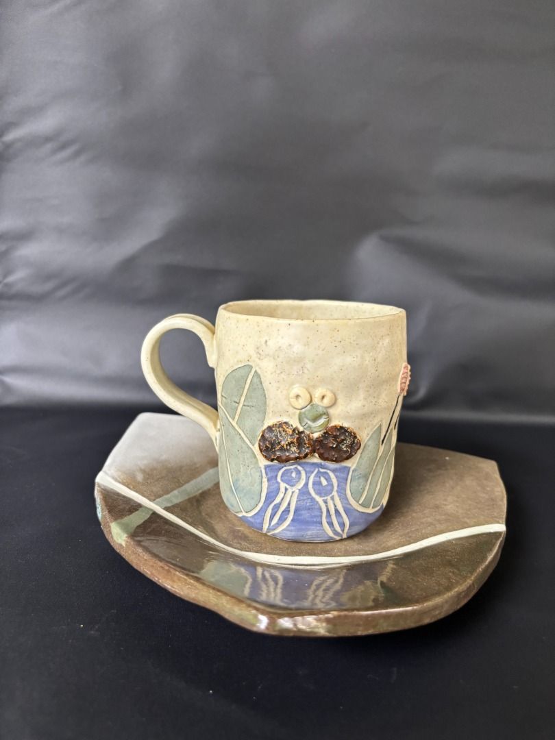 Handmade Ceramic Mug and Triangular Saucer Set (Category: Coffee & Tea Tableware) located in Baao. Submitted by apr1979 (Listing ID 1468785005, Image 2). Description: - Cup: 3.1 x 3.7 inches Triangle Footed Saucer/Trinket: 8 x 1.1 inches - Handmade ceramic mug with a cute raised frog face and plant designs - Glazed in charming beige, blue, and green tones - Comes with a unique triangular saucer featuring earthy brown and grey glazes with painted 
