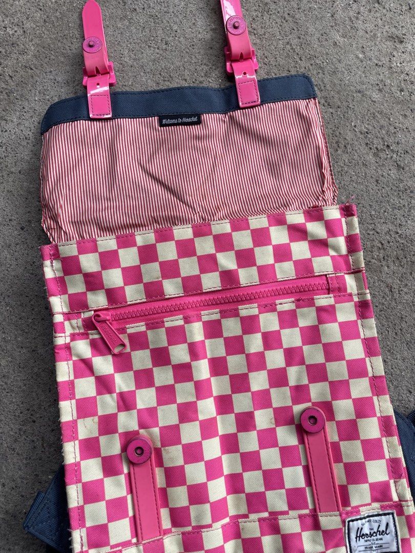 Herschel pink and white checkered backpack (Category: Backpacks) located in Polomolok. Submitted by madagascarts (Listing ID 1468798070, Image 2). Description: - Brand: Herschel - Design: Pink and white checkerboard pattern with dark grey flap and back straps - Pockets: Front zip pocket with stylish pink rubber straps and buckle closures - Interior: Signature red-and-white striped fabric lining - Details: Classic woven Herschel logo patch on the 