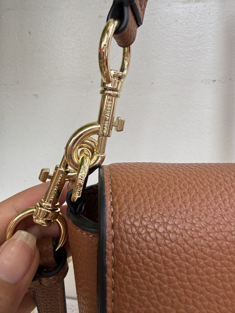 Marc Jacobs Brown Crossbody Bag with Gold Hardware (Category: Luxury Bags & Wallets) located in Consolacion. Submitted by naome1. (Listing ID 1468880107, Image 3). Description: Brown Marc Jacobs crossbody bag in textured leather. Bold white lettering on the front flap. Gold-tone hardware clips on the straps, all stamped with the brand name. Black edge painting along the seams. Includes both a short shoulder strap and a longer adjustable strap. Cl