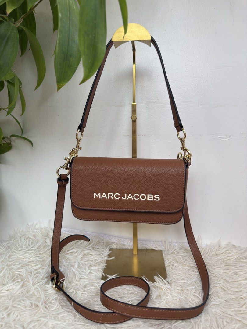 Marc Jacobs Brown Crossbody Bag with Gold Hardware (Category: Luxury Bags & Wallets) located in Consolacion. Submitted by naome1. (Listing ID 1468880107, Image 1). Description: Brown Marc Jacobs crossbody bag in textured leather. Bold white lettering on the front flap. Gold-tone hardware clips on the straps, all stamped with the brand name. Black edge painting along the seams. Includes both a short shoulder strap and a longer adjustable strap. Cl