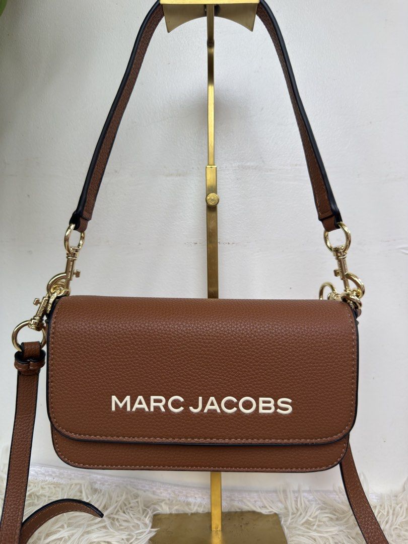 Marc Jacobs Brown Crossbody Bag with Gold Hardware (Category: Luxury Bags & Wallets) located in Consolacion. Submitted by naome1. (Listing ID 1468880107, Image 2). Description: Brown Marc Jacobs crossbody bag in textured leather. Bold white lettering on the front flap. Gold-tone hardware clips on the straps, all stamped with the brand name. Black edge painting along the seams. Includes both a short shoulder strap and a longer adjustable strap. Cl