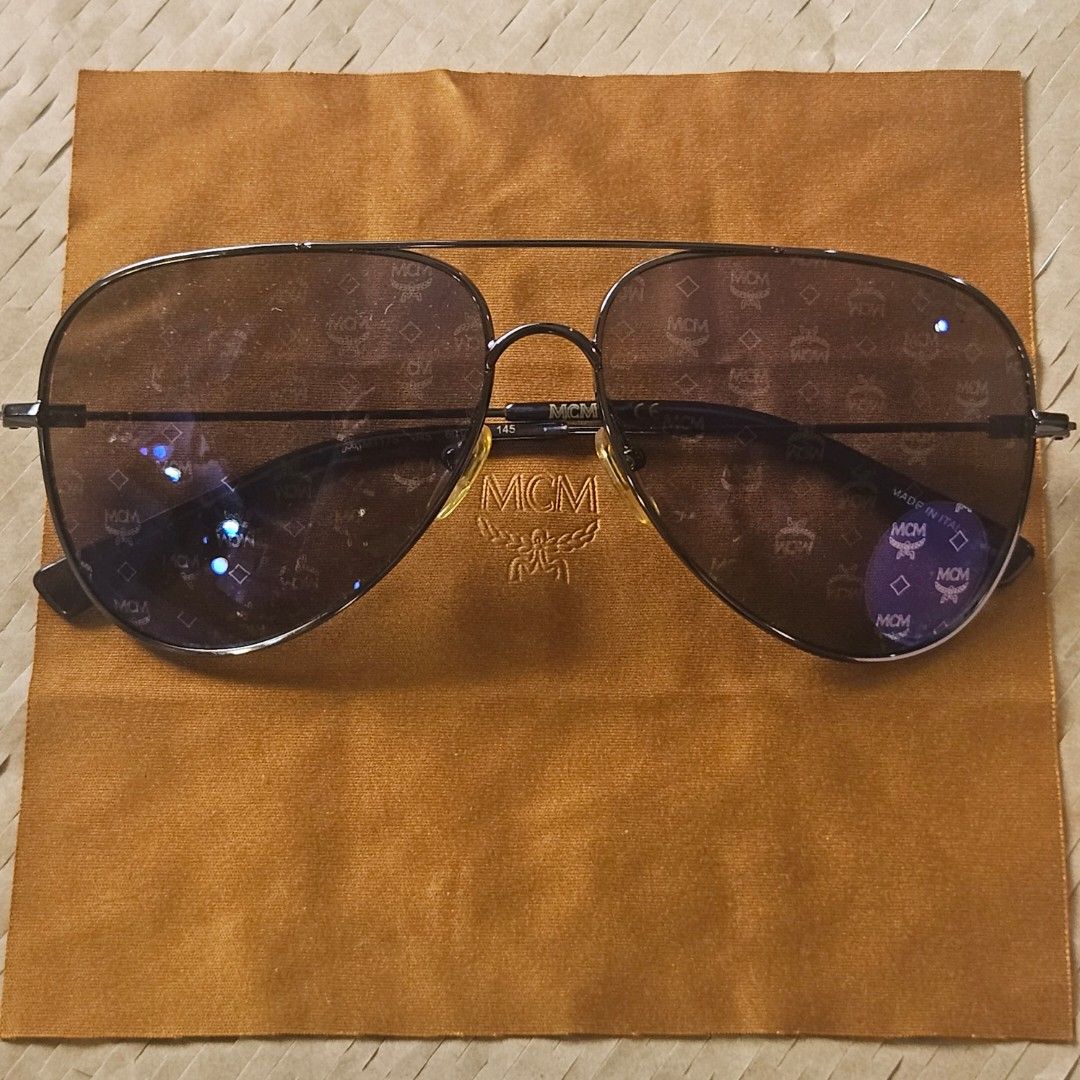 MCM Aviator Sunglasses (Category: Sunglasses & Eyewear) located in Imus. Submitted by a.niko (Listing ID 1468754356, Image 2). Description: - Brand: MCM - Style: Classic aviator / pilot shape with double brow bar - Lenses: Dark tinted with all-over MCM monogram pattern, clean condition - Frame: Sturdy metal frame - Origin: Made in Italy - Temple marking: MCM 145 - Inclusions: Comes with original MCM branded brown cloth pouch - Condition: Excellen