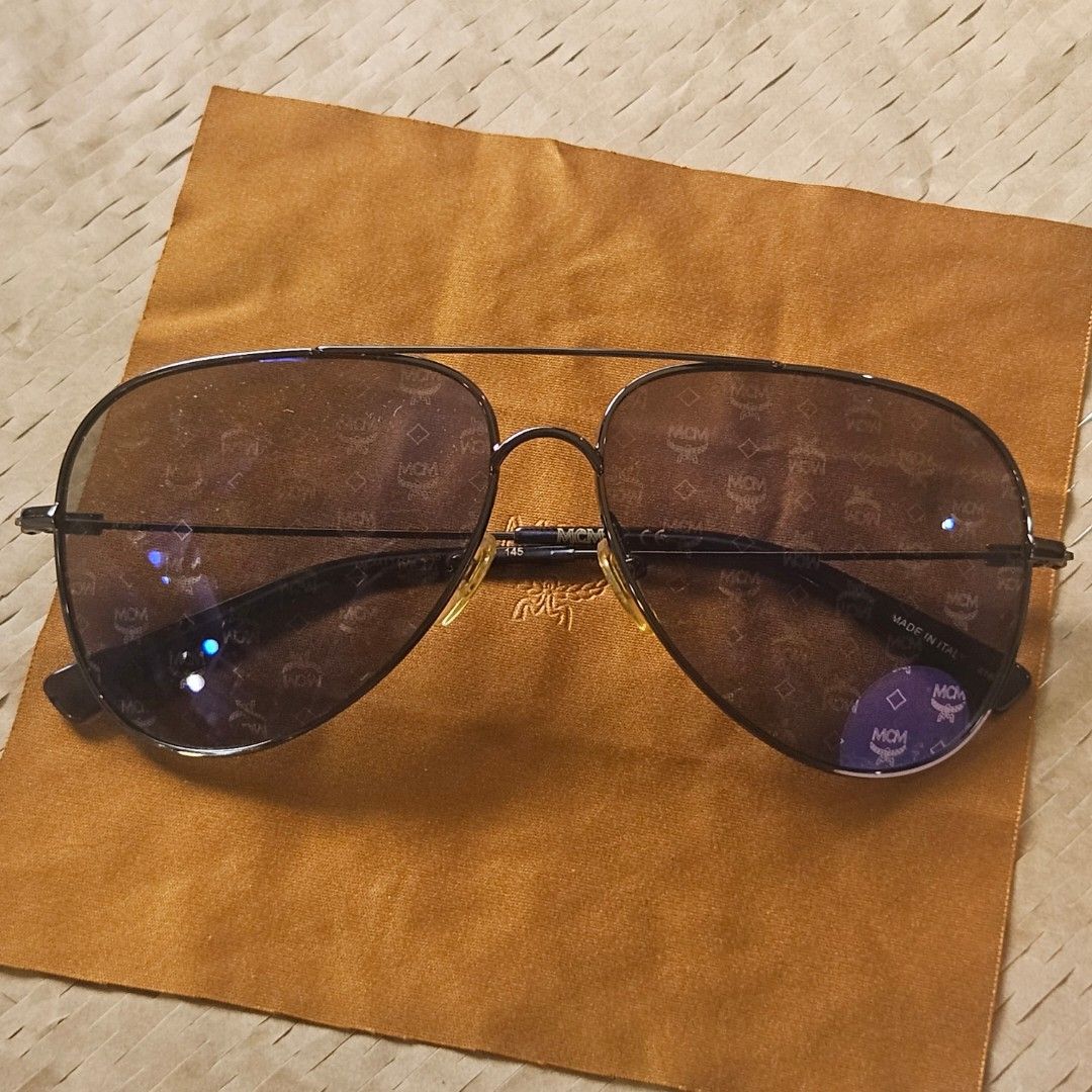 MCM Aviator Sunglasses (Category: Sunglasses & Eyewear) located in Imus. Submitted by a.niko (Listing ID 1468754356, Image 3). Description: - Brand: MCM - Style: Classic aviator / pilot shape with double brow bar - Lenses: Dark tinted with all-over MCM monogram pattern, clean condition - Frame: Sturdy metal frame - Origin: Made in Italy - Temple marking: MCM 145 - Inclusions: Comes with original MCM branded brown cloth pouch - Condition: Excellen