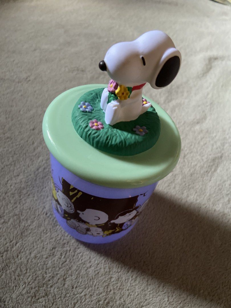 Peanuts Snoopy Mascot Jar with Figurine Lid (Category: Character Goods) located in Davao. Submitted by mengsmixitems (Listing ID 1468713007, Image 3). Description: - Official Peanuts merchandise imported from Japan - Purple plastic storage jar with cute character prints - Green lid featuring a 3D Snoopy figurine sitting on grass and flowers - Empty container only (treats not included) - Clean interior, perfect for desk or shelf organization.