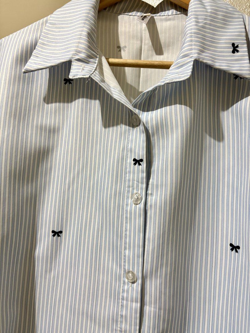 Blue and white striped button-down shirt with black bows (Category: Shirts) located in Caloocan City. Submitted by stella_mari27 (Listing ID 1469098861, Image 2). Description: - Style: Cropped long-sleeve button-down shirt - Design: Blue and white stripes with all-over black embroidered bow prints (even on collar and cuffs) - Fit: Regular fit with standard lapel collar and front button closure - Perfect for casual chic or preppy looks!.